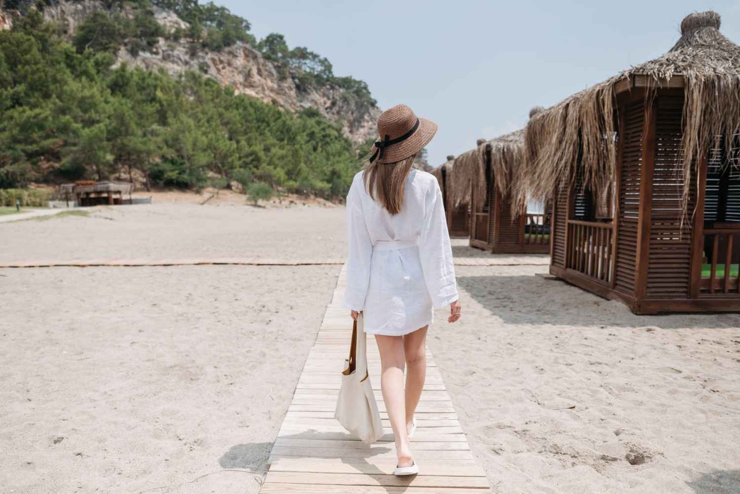 A woman in a white dress walks on a beach boardwalk surrounded by thatched cabanas, enjoying a summer day.